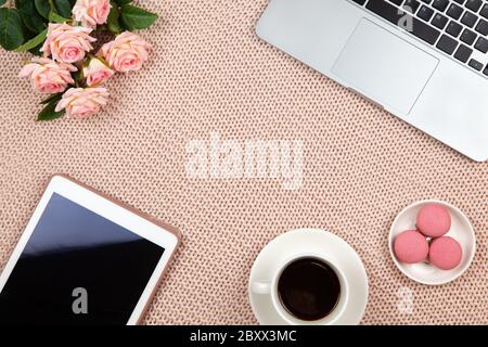 Concetto di lavoro da casa. Moderno spazio lavoro femminile, vista dall'alto. Laptop, caffè, torte, rose, tavoletta su coperta a maglia, spazio per la copia, piatto. Desktop Foto Stock