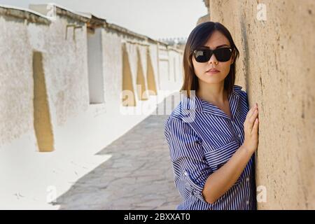 Piano medio ritratto di una bella giovane donna asiatica di fronte a un antico muro di argilla di strada in rovina. Tour della strada della seta dell'asia centrale Foto Stock