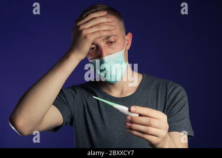 Uomo malato caucasico in una maschera di garza medica con palmo sulla testa tenendo termometro in altra mano, concetto di febbre. Foto Stock