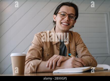 Bell'uomo che lavora freelance progetto, strategia di pianificazione, seduta sul posto di lavoro. Ritratto di giovane scrittore elegante scrivere note, guardando la fotocamera Foto Stock