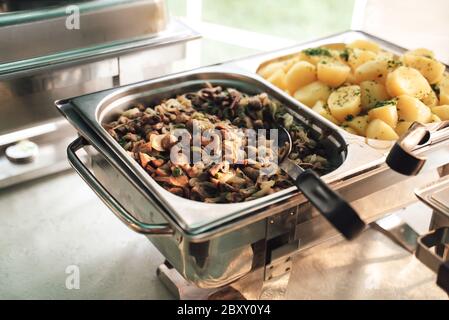 Due teglie quadrate in acciaio inossidabile sugli scaldabiberon. Patate cotte e funghi stufati come contorni. Tavolo a buffet self-service. Festa, festa Foto Stock