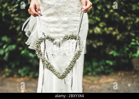 Sposa in piedi all'aperto e con decorazioni floreali a forma di cuore. Primo piano di cuore fiore su sfondo vestito bianco. Concetto di giorno di nozze. Foto Stock