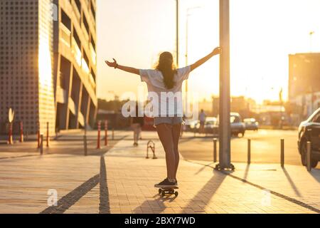 Ragazza su uno skateboard illuminato dai raggi del sole che tramonta. Corre su uno skateboard con le braccia sollevate, la schiena alla fotocamera. Messa a fuoco selettiva. Tramonto in città. Foto di alta qualità Foto Stock