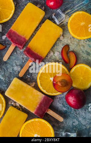 Primo piano di Possicles arancione e prugna su sfondo grigio, vista dall'alto. Popsicle di frutta / gelato a cubetti di ghiaccio, prugne fresche e pezzo d'arancia Foto Stock