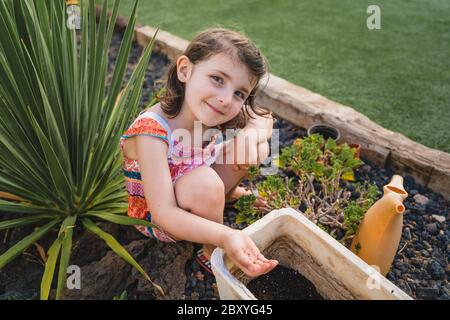 carino bambina nel giardino del cortile tiene semi di pianta in mano in pentola, sorridendo a macchina fotografica seduta giù sul terreno Foto Stock