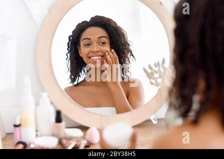 Donna nera graziosa che guarda nello specchio e applica la crema idratante facciale Foto Stock