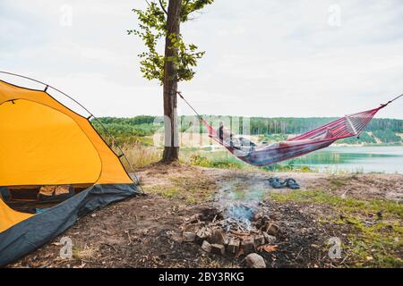 uomo che si posa su amaca sulla spiaggia del lago vicino al fuoco del campo Foto Stock