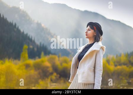 Bella donna in un cappotto bianco e cappello nero che posa su uno sfondo di montagne autunnali Foto Stock