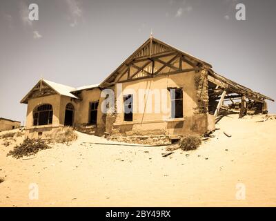 Edifici fantasma della vecchia città mineraria di diamanti Kolmanskop vicino a Luderitz in Namibia. Rovine abbandonate di casa affondate nelle dune di sabbia del deserto del Namib. Fotografia con tonalità vintage. Foto Stock