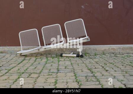 Fila di sedie in metallo bianco danneggiate con gamba rotta, sedie inclinate vengono abbandonate su pavimentazione in pietra nel parco Foto Stock