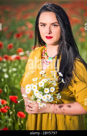 Ritratto di una bella giovane donna in una blusa gialla nel mezzo di un campo di papavero che tiene in mano un bouquet di fiori bianchi Foto Stock