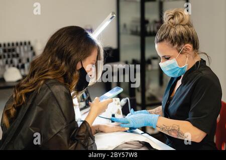 Beautician mascherato, dipingendo le unghie del suo cliente in un centro di bellezza, mentre registra un video con il suo smartphone. Foto Stock