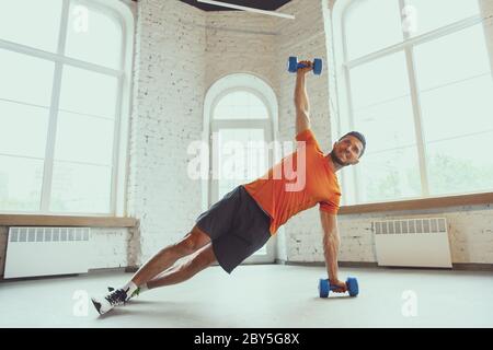 Manubri. Giovane uomo caucasico addestramento a casa durante la quarantena di epidemia di coronavirus, facendo esercizi di fitness, aerobica. Rimanere sportivi durante l'isolamento. Concetto di benessere, sport, movimento. Foto Stock