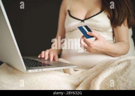 Donna pensiva shopping online con carta di credito e laptop, seduta su divano a casa. Giovane donna in attesa di rete da surf, acquisto di merci per neonato Foto Stock