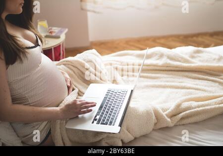 Bella donna incinta rilassante con il suo computer portatile mentre giaceva su un letto di casa Foto Stock