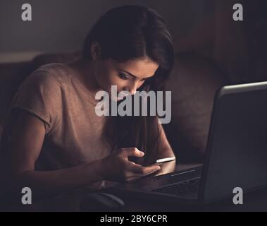 Sorridente giovane donna pensante seduta vicino al monitor del computer e testando sul telefono cellulare su sfondo scuro ombra casa. Poirtrait vintage in tonalità Foto Stock