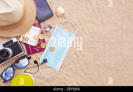 Alcuni oggetti da spiaggia, una maschera protettiva, un passaporto e alcuni contanti, uno smartphone, cappello, macchina fotografica e occhiali da sole sulla spiaggia di sabbia. Spazio per il testo. Concep Foto Stock