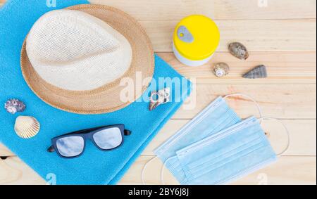 Vista dall'alto di alcuni oggetti della spiaggia come asciugamano, cappello e occhiali da sole insieme a un paio di maschere facciali di protezione su un pannello di legno. Concetto di vacanza estiva Foto Stock