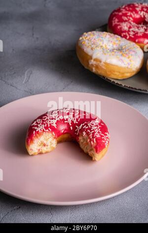 Ciambella rosa morso con spruzzini su piatto rosa vicino a ciambelle di cioccolato e vaniglia, dolce cibo da dessert smaltato su fondo testurizzato in cemento Foto Stock