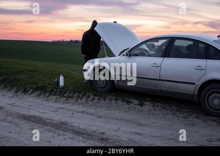 Uomo in piedi vicino all'auto con cofano aperto al tramonto. Guasto dell'auto. Uomo confuso si alza con cappuccio aperto con fuoco morbido selettivo. Foto Stock