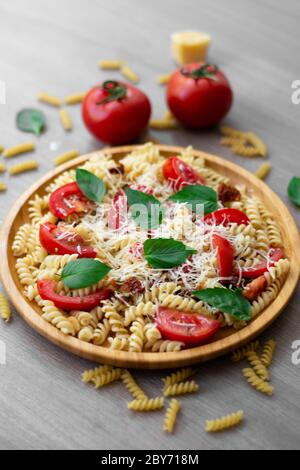 Insalata estiva di pasta fredda con fusilli, pomodori freschi, pomodori secchi, parmigiano grattugiato e foglie di basilico fresco verde in un piatto di legno Foto Stock