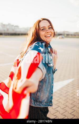 Giovane donna dai capelli rossi bella avvolta con bandiera nazionale degli stati uniti sopra il sole. Retroilluminazione morbida. Giorno dell'indipendenza, sogno americano, concetto di libertà Foto Stock