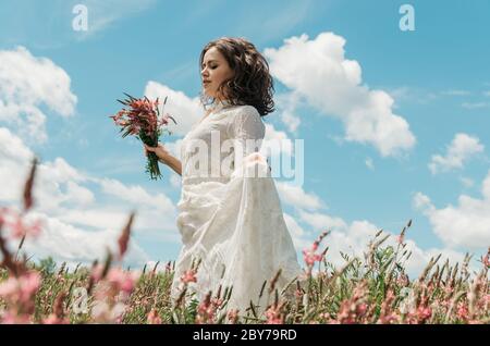 Ritratto di una giovane donna in un lungo vestito bianco che tiene un bouquet di fiori e il suo vestito mentre cammina in un campo di fiori contro il cielo blu Foto Stock