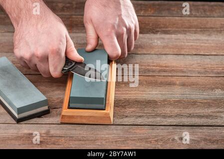 Uomo affilando il suo coltello da tasca con una pietra da ciminello su un rustico tavolo di legno Foto Stock