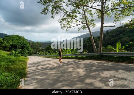 Donna in t-shirt nera che gode di vista tropicale foresta mentre si trova su una strada vuota. Montagne e nuvole bianche su un cielo blu. Vacanza tropicale estiva Foto Stock