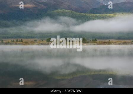 Vista magica con riflessi di montagne coperte di verde foresta, colline, alberi e bella nebbia nelle acque dolci del lago, all'inizio della mattina Foto Stock