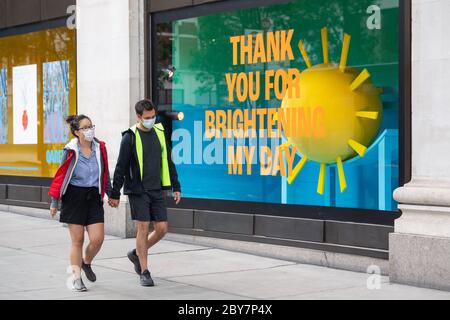 Due persone che indossano maschere facciali di protezione passano la finestra a Selfridge's su Oxford Street, Londra, con messaggi di ringraziamento da parte del personale di Selfridge a persone che li hanno aiutati durante il blocco del coronavirus. Foto Stock