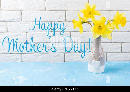 Biglietto di auguri per il giorno della mamma. Mazzo di fiori giallo fresco su tavolo blu davanti a muro di mattoni bianchi. Narcissus giallo o fiori di daffodil. Foto Stock