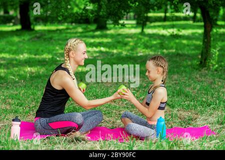 Madre e sua figlia in abbigliamento sportivo che tiene una mela verde e si siede sul tappeto in parco. Foto Stock