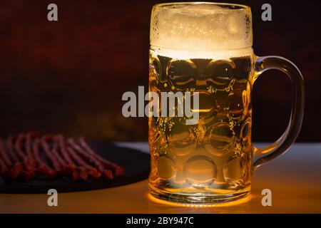 Un bicchiere pieno di birra con testa di schiuma in un pub servito su un bancone con salsiccia secca piccante per spuntini con spazio laterale Foto Stock