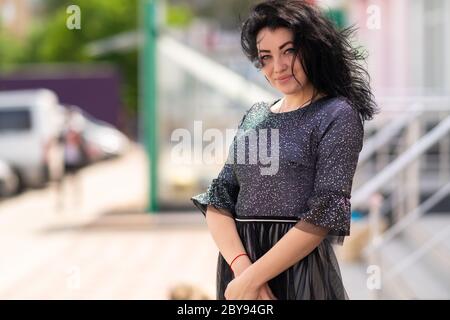 Donna felice e cordiale con un sorriso caldo e piacevole in piedi in città in una giornata ventosa con i suoi capelli che soffiano intorno al suo volto sorridente alla macchina fotografica Foto Stock