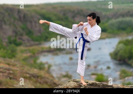 Giovane donna che pratica kickboxing all'aperto in natura che si bilancia su una roccia sopra un fiume nella valle sottostante Foto Stock