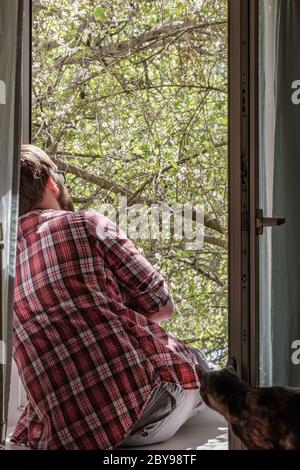 Uomo seduto su un davanzale alla finestra aperta e godendo i fiori di ciliegio vicino al suo gatto curioso guardare con interesse. Foto Stock