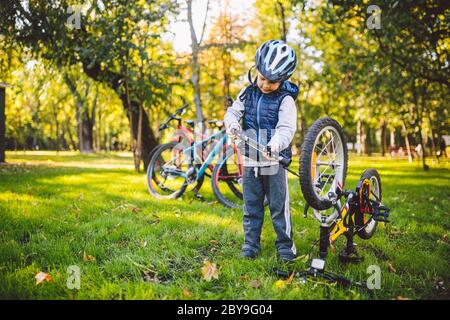 Il bambino caucasico in un casco impara a riparare la sua moto. Il ciclista dei bambini controlla il meccanismo di una bicicletta in una radura di erba verde nel parco Foto Stock