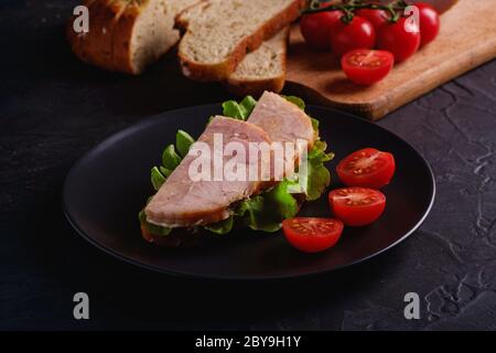 Sandwich con carne di prosciutto di tacchino, insalata verde e pomodori ciliegini freschi a fette su piatto nero vicino agli ingredienti sul tagliere Foto Stock