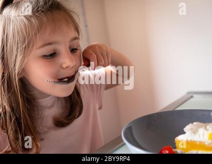 Bella bambina godendo un boccaglio di torta mangiare fuori del cucchiaio come lei si siede a tavola in un ritratto ravvicinato Foto Stock