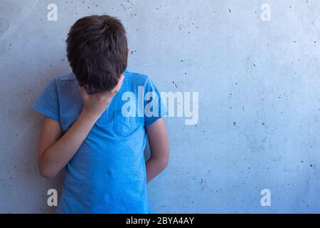 Il ragazzo triste si alzò in piedi e si appoggiò su una parete grigia. Difficoltà di apprendimento, problemi familiari, bullismo, depressione, stress o concetto di frustrazione Foto Stock