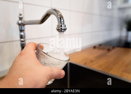 acqua potabile e sicura da bere. uomo che riempie un bicchiere d'acqua da un rubinetto in acciaio inox. mano maschile che versa l'acqua nel bicchiere da cromo Foto Stock