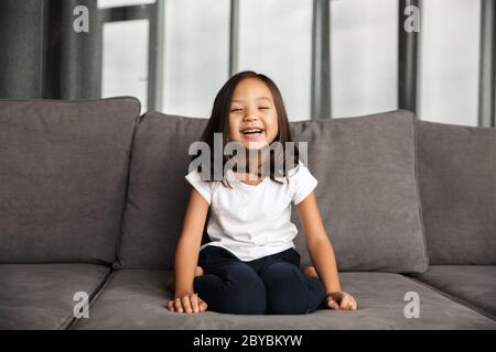 Foto di gioiosa bambina asiatica che indossa abiti casual e ride mentre si siede sul divano a casa Foto Stock