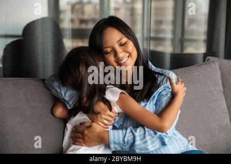 Foto di donna asiatica in gravidanza con la sua piccola figlia che si agguenta e sorride mentre si siede sul divano a casa Foto Stock