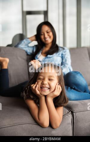 Foto di felice donna asiatica incinta con la sua piccola figlia che ride mentre si riposa sul divano a casa Foto Stock