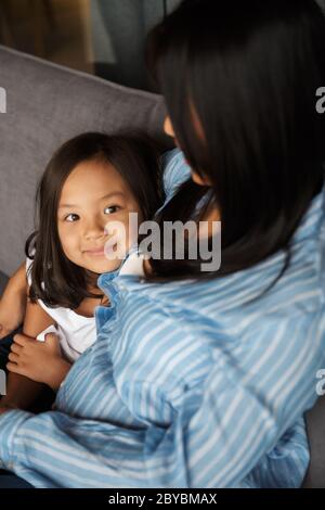 Foto di giovane donna asiatica incinta con la sua piccola figlia che si agguanava mentre si siede sul divano a casa Foto Stock