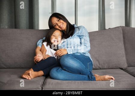 Foto di donna asiatica in gravidanza con la sua piccola figlia che si agguenta e sorride mentre si siede sul divano a casa Foto Stock