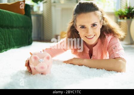 Ritratto di giovane donna sorridente in blusa e pantaloni bianchi con banco di piggy mentre si posa su tappeto bianco a casa moderna in giornata di sole. Foto Stock