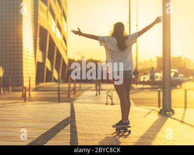 Ragazza su uno skateboard illuminato dai raggi del sole che tramonta, con le braccia sollevate in alto, la schiena alla fotocamera. Messa a fuoco selettiva. Foto Stock