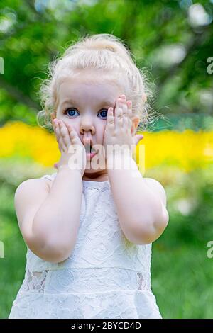 la bambina con i capelli biondi in un vestito bianco tiene le mani alle guance e mostra sorpresa Foto Stock